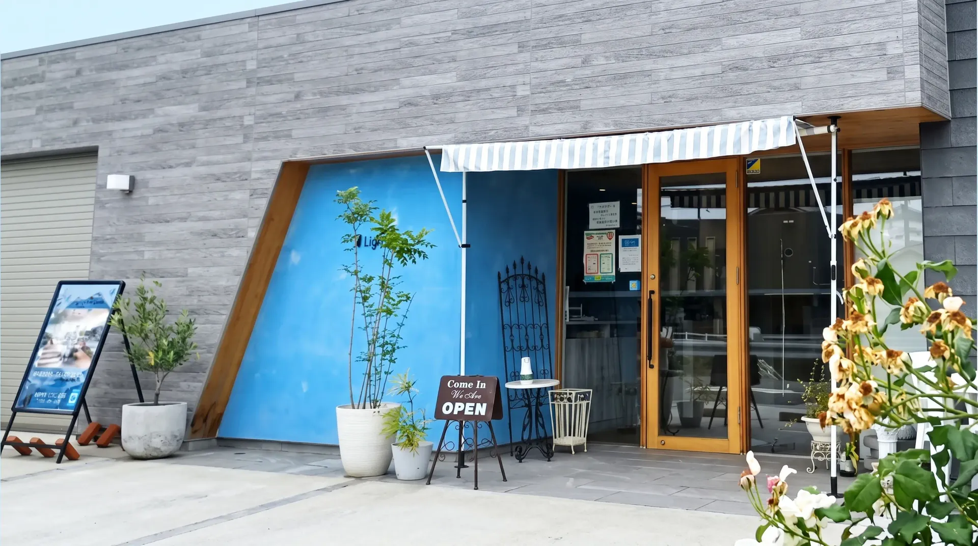 カフェ ライトブルー の外観。水色のファサードに木の扉、OPEN の看板と鉢植えを置いた入口